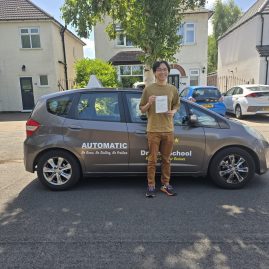 A smiling young man stands beside a grey car with "AUTOMATIC - No Gears, No Stalling, No Problem" and "Driving School" written on the side. He is holding up what appears to be a driving test pass certificate. The scene is on a residential street with houses and parked cars in the background.
