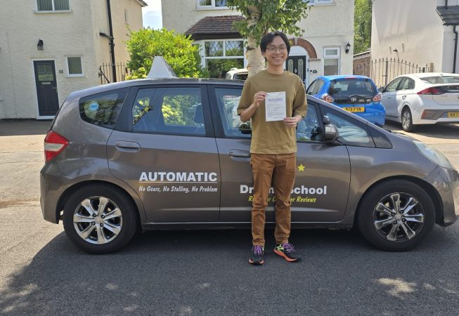 A smiling young man stands beside a grey car with "AUTOMATIC - No Gears, No Stalling, No Problem" and "Driving School" written on the side. He is holding up what appears to be a driving test pass certificate. The scene is on a residential street with houses and parked cars in the background.